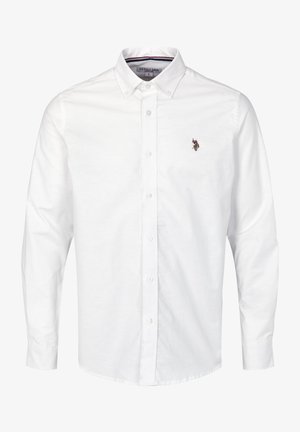 Homme portant une chemise blanche boutonnée à manches longues, un pantalon sombre ajusté et des chaussures habillées noires, debout devant un fond uni.