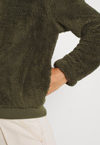 Oliwkowy polar z miękką, teksturowaną powierzchnią. Posiada prążkowane mankiety i pas. Dopasowane rękawy oraz luźny krój.