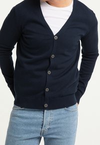 Mörkblå kofta i mjukt tyg med V-ringning, sex knappar framtill, ribbade mudd och fåll, paired med en vit T-shirt under.