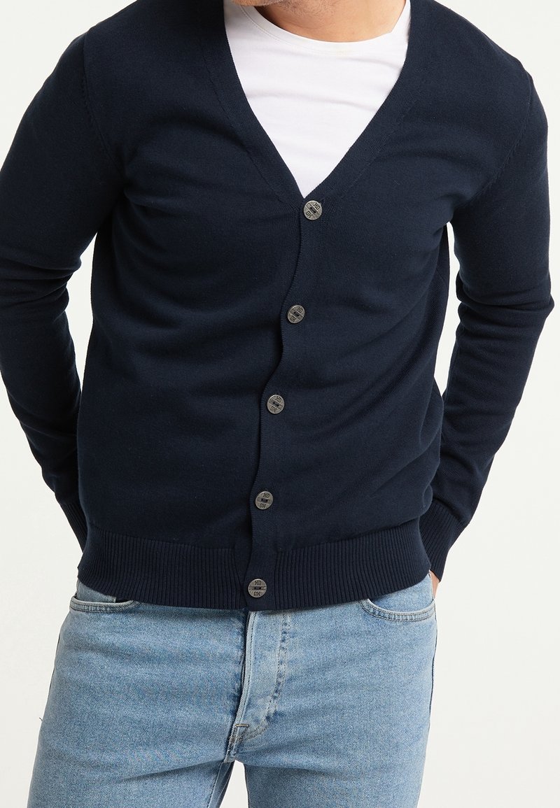 Mörkblå kofta i mjukt tyg med V-ringning, sex knappar framtill, ribbade mudd och fåll, paired med en vit T-shirt under.