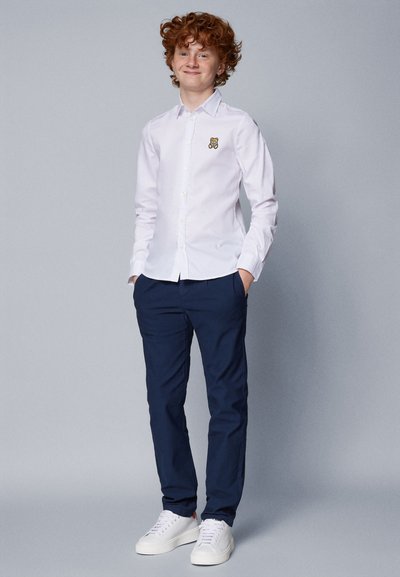 Camicia bianca con bottoni caratterizzata da un logo di un orso sul lato sinistro del petto, abbinata a pantaloni blu navy e sneakers bianche. Sfondo semplice.