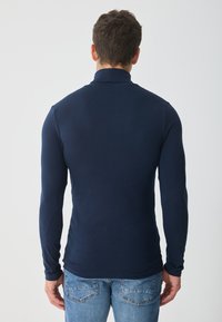 Polo bleu închis cu mâneci lungi, realizat dintr-un material neted, având un guler înalt și un design mulat, asortat cu blugi albastru deschis.