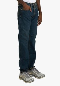 Jeans di denim blu con taglio a gamba dritta, cinque tasche e una leggera sbiaditura, abbinati a scarpe da corsa grigie con pannelli in mesh e lacci.