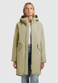 Jeune femme portant une parka à capuche beige clair avec des poches à rabat, une chemise blanche et un jean bleu, posant devant un fond uni.
