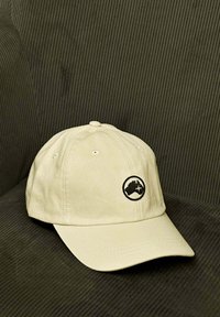 Gorra de béisbol de algodón beige con una corona redondeada, visera curvada y un logotipo circular negro que presenta un mapa estilizado y un avión.