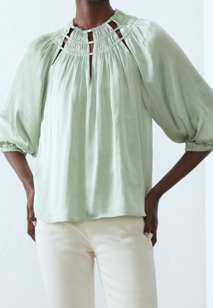 Woman wearing a light green blouse with gathered neckline and cutout details, paired with off-white pants, standing with hands on hips.