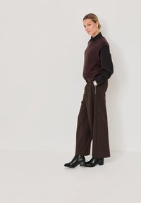 Chaleco de lana marrón sobre una camisa negra, combinado con pantalones anchos marrones y botas negras hasta el tobillo. El conjunto presenta líneas limpias y un acabado texturizado.