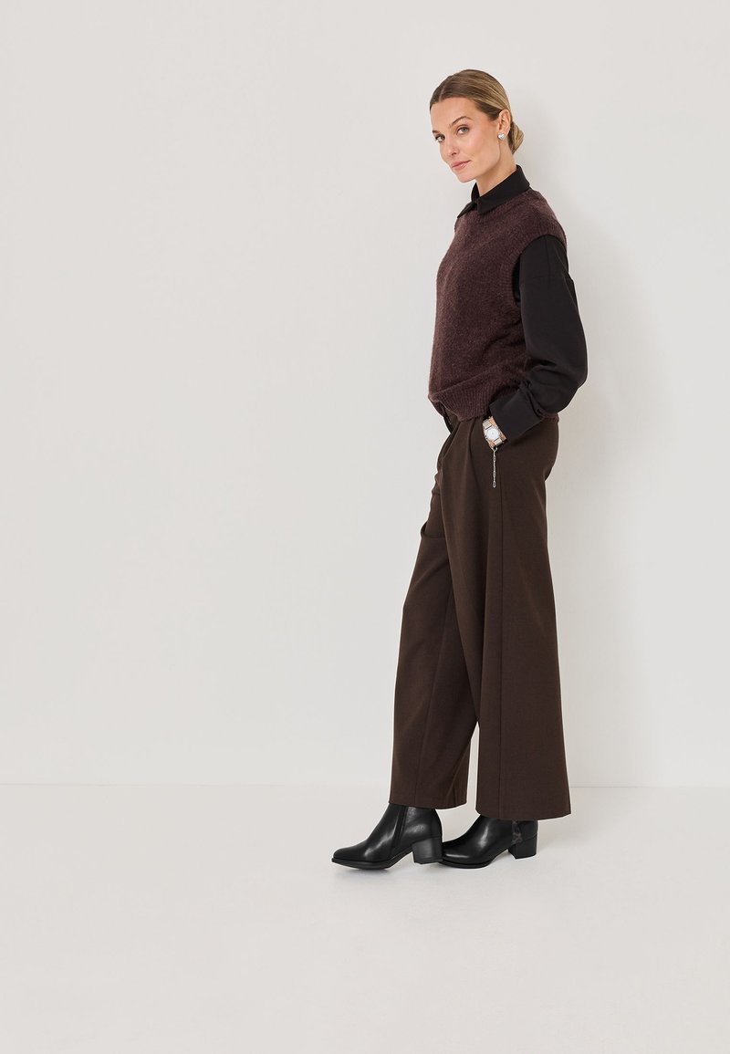 Chaleco de lana marrón sobre una camisa negra, combinado con pantalones anchos marrones y botas negras hasta el tobillo. El conjunto presenta líneas limpias y un acabado texturizado.