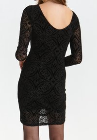 Frau in einem figurbetonten schwarzen Kleid mit einem durchsichtigen gemusterten Überwurf und langen Ärmeln, von hinten vor einem schlichten Hintergrund gezeigt.