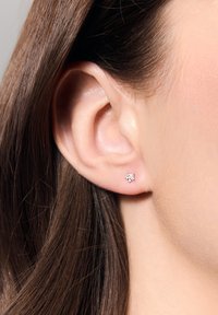 Srebrny kolczyk w kształcie gwiazdki, ozdobiony przezroczystymi kamieniami, przymocowany do ucha modelki z prostymi brązowymi włosami. Gładkie tło skóry.