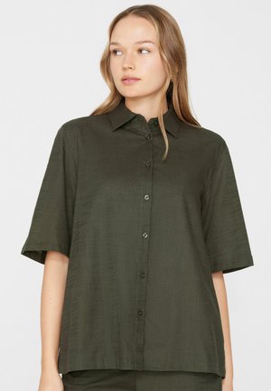 Junge Frau schaut seitlich, trägt ein locker sitzendes, dunkelgrünes, kurzärmeliges Hemd mit Knöpfen vor weißem Hintergrund.