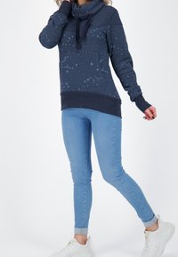 Sudadera de marina con cuello tipo cowl, que presenta un patrón de pájaros, bolsillo frontal y puños acanalados. Combinada con jeans ajustados de color azul claro y zapatillas blancas.