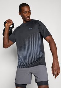 T-shirt de sport noir avec motif à rayures bleues, manches courtes et logo. Short de sport gris avec fentes sur les côtés. Tenant un kettlebell noir.