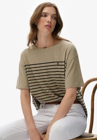 Beige stribet T-shirt med løs pasform, sorte vandrette striber, rund halsudskæring og et lille logo på brystet, parret med hvide bukser.