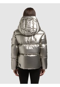 Donna che indossa una giacca a vento argentata metallizzata con cappuccio con la scritta "KHUN", vista di spalle su uno sfondo neutro.