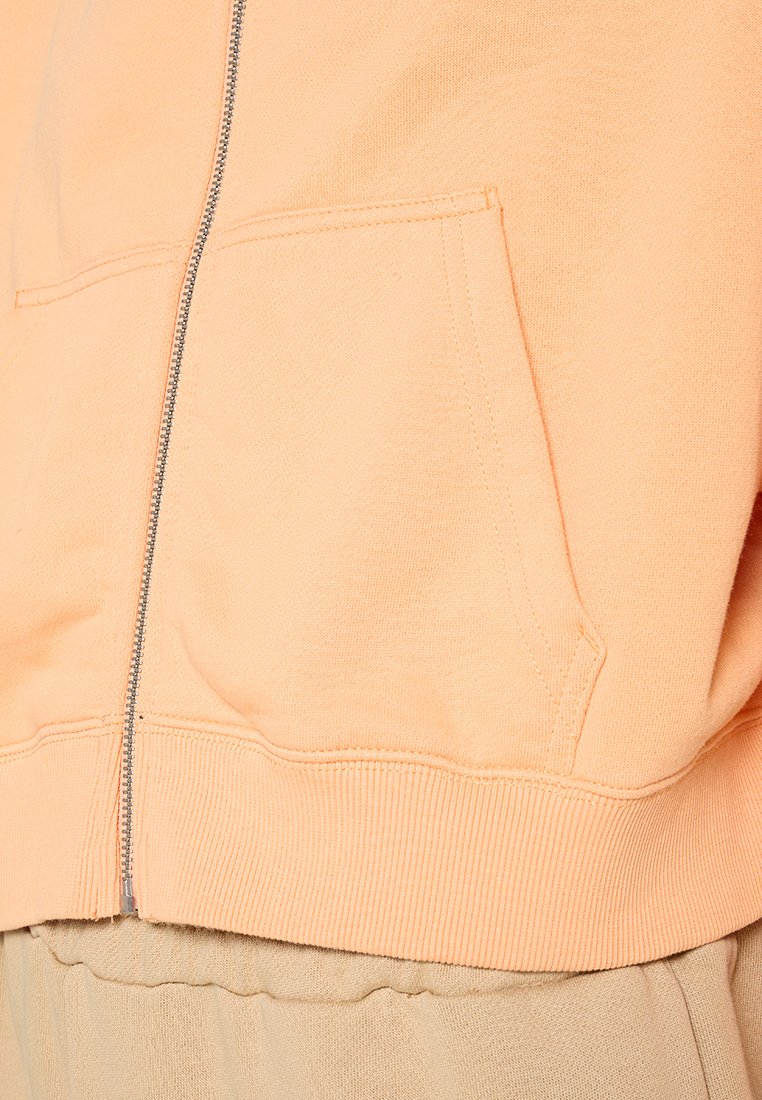 Pfirsichfarbene Zip-Hoodie mit einer glatten Baumwolloberfläche. Verfügt über Fronttaschen und gerippte Bündchen. Darunter ist ein spielerischer Jumpsuit in Beige sichtbar.