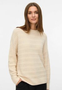 Jeune femme aux longs cheveux bruns portant un pull en tricot beige à motifs et un pantalon noir, se tenant avec une main dans la poche devant un fond blanc.