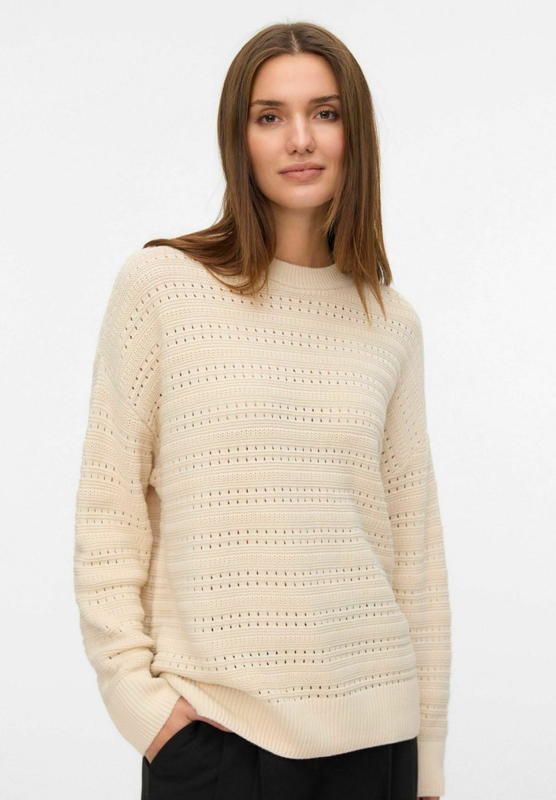Jeune femme aux longs cheveux bruns portant un pull en tricot beige à motifs et un pantalon noir, se tenant avec une main dans la poche devant un fond blanc.
