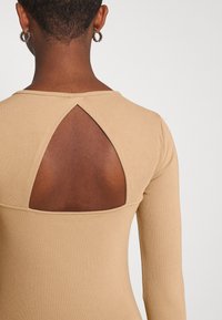 Maglietta beige a maniche lunghe con scollatura rotonda e dettagli di schiena aperta a forma triangolare. Texture liscia, design aderente e stile minimalista.