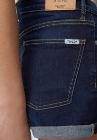 Ciemnoniebieskie dżinsowe szorty z podwiniętymi mankietami, z naszywką ze skóry i małą metką materiałową. Szycie w kontrastowym żółtym kolorze.