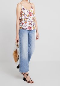 Femme portant un haut sans manches à fleurs, un pantalon large bleu clair, des talons noirs à lanières, tenant un sac à main transparent avec une pochette marron à l'intérieur.