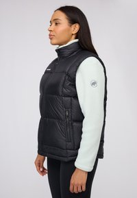 Schwarze Steppweste mit hohem Kragen, gestepptem Design und Reißverschlusstaschen, getragen über einem hellgrünen Fleecepullover mit Logo am Ärmel.