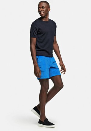 Uomo sorridente che cammina, indossa una camicia nera a maniche corte, pantaloncini blu brillanti e scarpe slip-on nere con suola bianca.