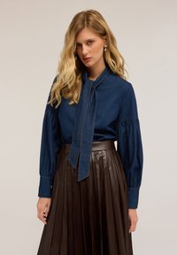 Blusa di denim con colletto annodato e maniche a sbuffo lunghe, abbinata a una gonna plissettata in pelle marrone. Texture liscia con cuciture a contrasto.