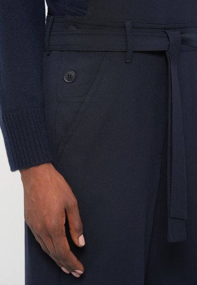 Temno modri hlače s prilegajočim dizajnom, ki imajo zavezan pas, dve sprednji žepi in gumbe kot detajle. Mehka tekstura tkanine.