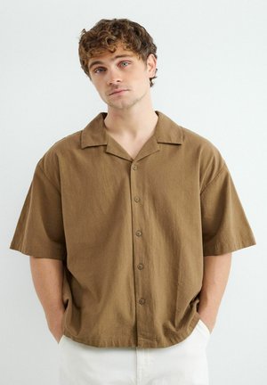 Mladý muž kučeravými vlasmi, oblečený v voľnej hnedej krátkorukávovej košeli s gombíkmi a bielych nohaviciach, ruky v vreckách, stojaci na bielom pozadí.