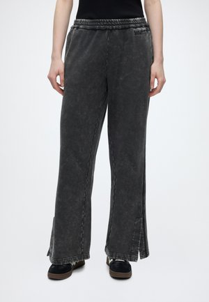 Personne portant un pantalon ample gris foncé délavé à jambes larges avec des fentes en bas et des baskets noires avec des lacets blancs.