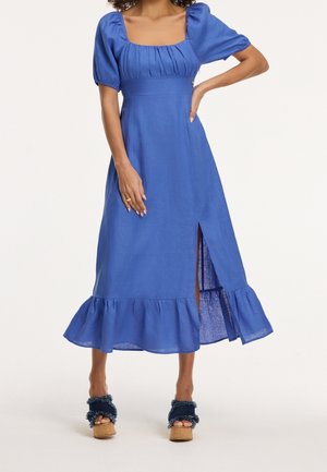 Vrouw die een blauw midi-jurk draagt met pofmouwen, een vierkante halslijn, een voorsplit, een gerimpelde zoom en plateausandalen, tegen een effen achtergrond.