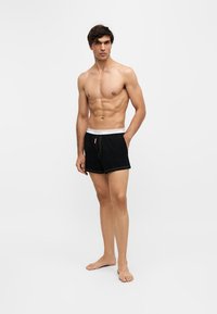 Jeune homme debout pieds nus portant un boxer noir avec une ceinture blanche sur un fond clair uni.