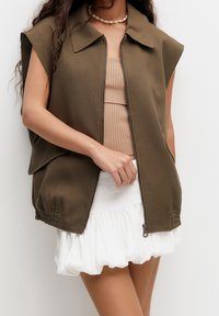 Gilet sans manches vert olive avec une fermeture éclair à l'avant, grand col et détail de ourlet élastique, associé à un haut écru à côtes et à une jupe blanche à volants.