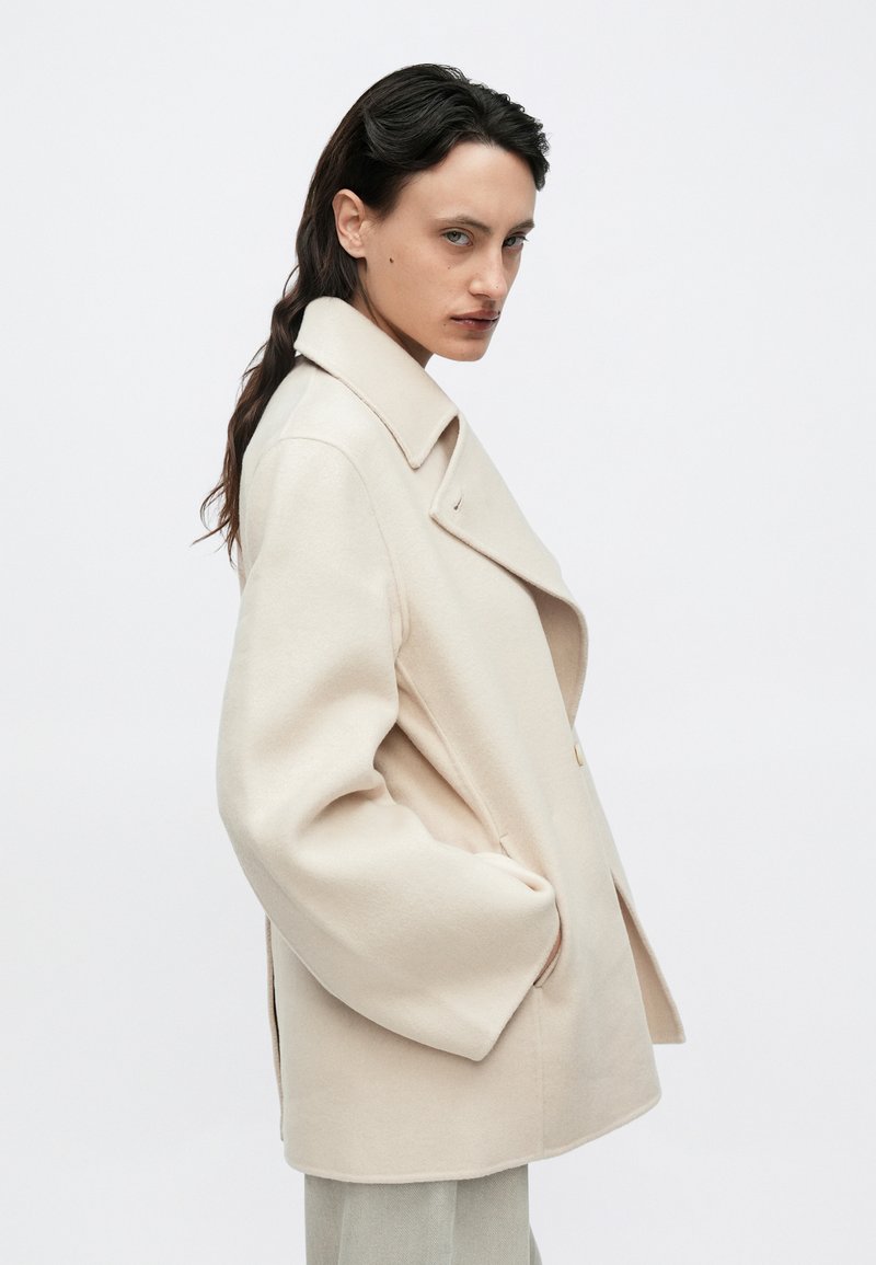 Beige double-breasted coat with a notched collar, side pockets, and a relaxed fit. Soft fabric with a smooth texture and minimal design.