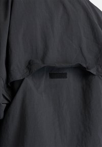 Giacca nera leggera con tessuto liscio e strutturato, colletto parzialmente aperto e chiusura con velcro.