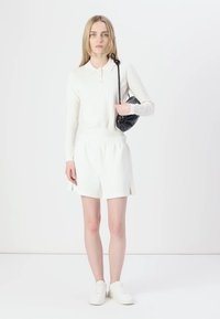 Polo de manga larga blanca con texture acanalada, combinado con pantalones cortos blancos con aberturas laterales. La modelo sostiene un bolso negro de forma redonda y lleva zapatillas blancas.