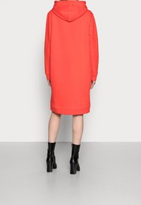 Hoodie klänning i ljus korall, med lång ärm och en nederkant som når mitt i vadder, kompletterad med svarta ankelskor.