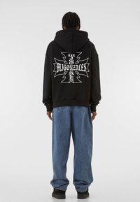 Black hooded sweatshirt with white graphic design on the back, paired with loose-fitting blue denim jeans and black sneakers.
