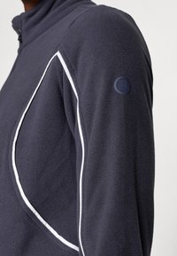 Fleecejacke in Dunkelblau mit weißen Paspeln. Merkmale sind ein hoher Kragen und ein kleines Logo-Detail am linken Ärmel. Weiche Textur.