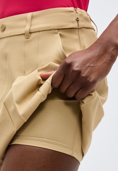 Hånd trækker i beige ydershortsstof for at afsløre tætsiddende indershorts på mørk hud Farvet ben iført rød top.