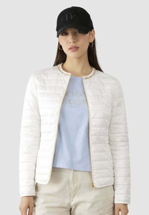 Donna che indossa una giacca trapuntata bianca, una t-shirt azzurra chiara stampata, pantaloni beige, un cappello nero e orecchini a cerchio su uno sfondo grigio.