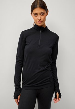 Vrouw draagt een zwart long-sleeve top met halve rits en duimgaten, staande tegen een effen lichtgrijze achtergrond.
