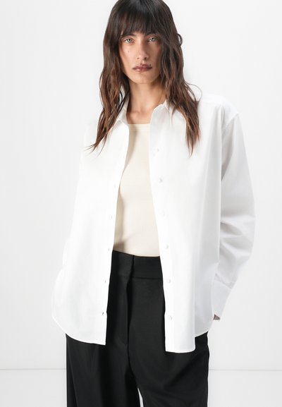 White button-up shirt with a relaxed fit, long sleeves, and rounded hem. Worn over a beige top and paired with black trousers.