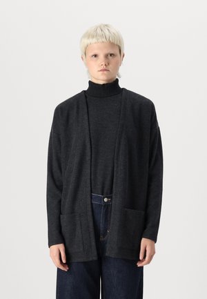 Mørk grå strikkegenser med åpen front, lange ermer og to frontlommer, båret over en svart turtleneck og blå jeans.