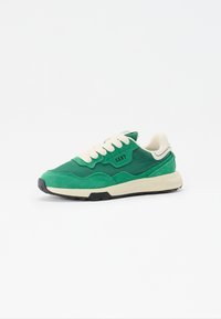 Green sneaker with a mix of suede and synthetic materials, cream laces, thick white midsole, and black rubber outsole. Brand logo on the side.