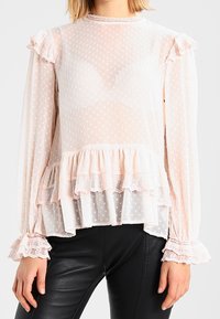 Jasnoróżowa, przezroczysta bluzka z kropkowanym wzorem, z falbaniastymi mankietami i warstwowym dołem. Posiada koronkowe wykończenie i długie rękawy, zestawiona z czarnymi skórzanymi spodniami.