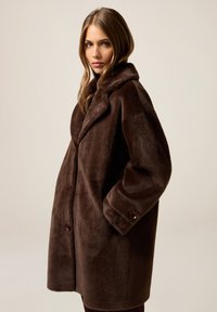 Kobieta o długich włosach, ubrana w zapinany na guziki, ciemnobrązowy, pluszowy płaszcz z szerokimi klapami, z rękami w kieszeniach, stojąca na jednolitym tle.