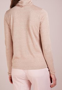 Růžovozlatý dlouhý rukáv turtleneck s jemnou třpytivou strukturou, přiléhavý střih a vysoký límec. Pozadí v jemně růžové barvě.