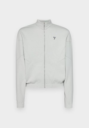 Veste zippée cropped gris clair avec col haut, en tissu texturé, coutures latérales et un petit logo sur la zone de la poitrine gauche.